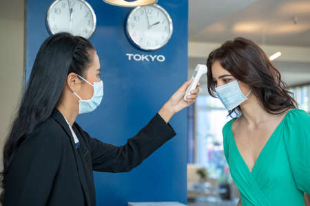 Check-in at the hotel.Reception wear protective face mask use infrared thermometer checker or temperature gun on hotel guest,Lifestyle new normal after  virus.の写真素材