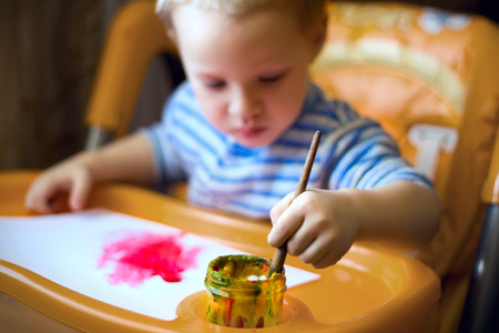 A little boy is sitting at the kids table, holding a brush, paint, paints.の写真素材
