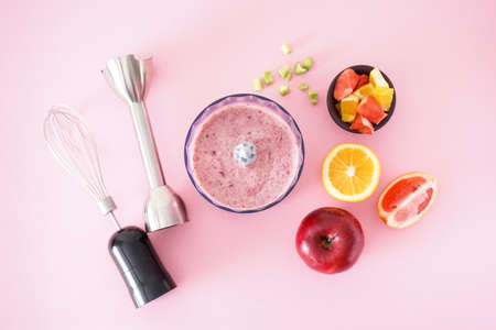 Black plastic electric hand blender and accessories with fruits and smoothie ready on pink background, flat lay.の写真素材