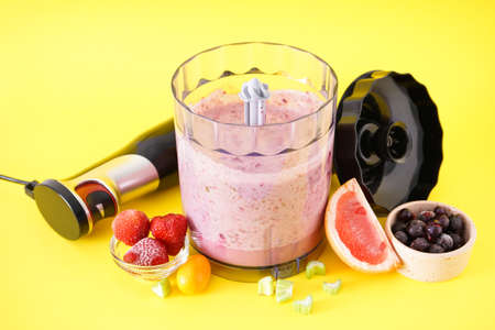 Black plastic electric hand blender and accessories with fruits and smoothie on a yellow background.の写真素材