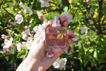 Perfume in the hand of a young woman on a green background, close-up. Blossoming apple orchard in...