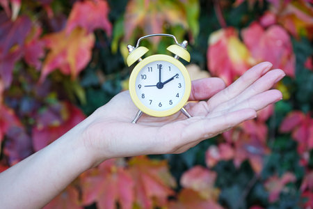 Reminder to switch to winter time. A woman holds an alarm clock against the background of autumn...