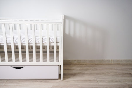 A clean, modern white baby crib with a mattress, placed in a bright minimalist nursery room with wooden flooring and sheer curtains. The crib has a lower storage drawer, ideal for baby essentials.の写真素材