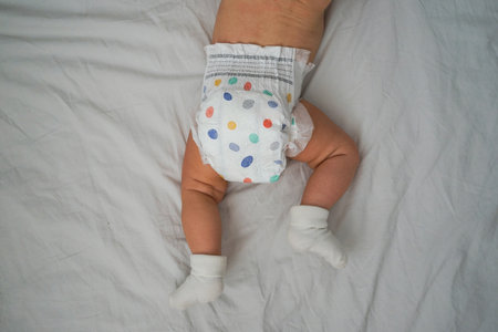 Top view of a baby wearing a diaper with colorful dots and white socks, lying on a white sheet....