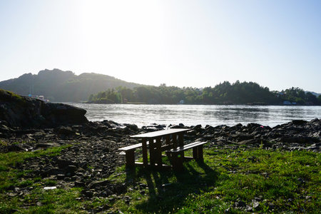 A serene early morning view of a wooden picnic table by the calm rocky shoreline, with sunlight...