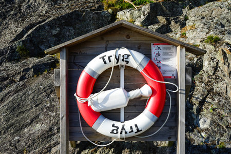 Detailed close-up of a red and white lifebuoy with rope mounted on a wooden rescue station along a rocky coastal cliff.の写真素材