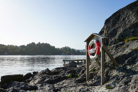 A bright lifebuoy station mounted on rocky coastal terrain, overlooking calm water and surrounded by...