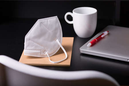 Close up view of a black work desk interior with a laptop computer, a cup of coffee, paper, pen, plant, face mask for coronavirus black backgroundの写真素材