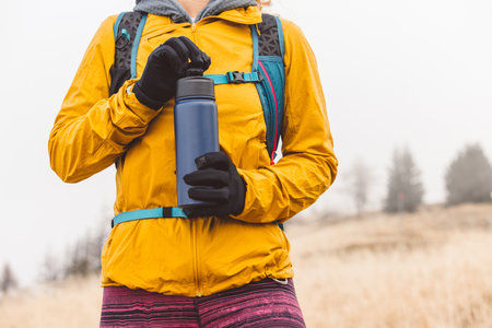 Unrecognizable mountaineer waist up in yellow jacket holding a blue thermo flask, up in the...
