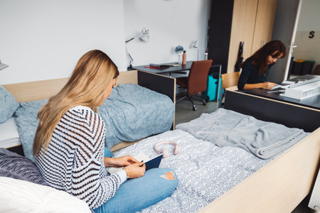 One woman student sitting on the bed reading a book and the other sitting by the desk working on a...