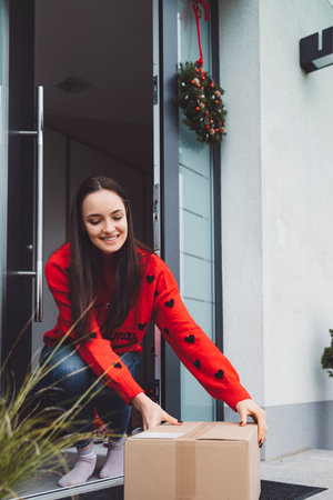 Smiling young woman picking up a delivery box from her front door, wearing a red Christmas sweaterの写真素材