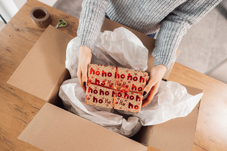 Top down view woman hands holding a Christmas present, putting it inside a cardboard box, ready to send in the mailの写真素材