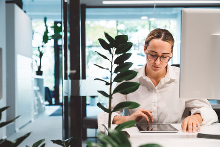 Young blonde woman standing behind a green plant looking at something on her digital tabletの写真素材