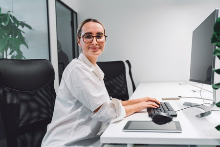 Side portrait of smiling businesswoman with glasses sitting behind her computer in the officeの写真素材