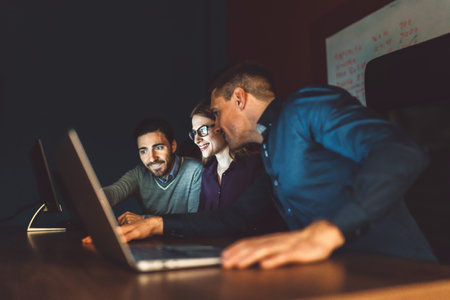 Group of friends starting up a company working late hours, smiling at computer screen at nightの写真素材
