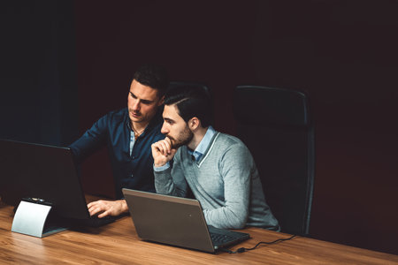 Two business man working in a dark room, on laptop and computer, thinking about ideas for a new campaignの写真素材