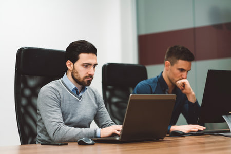 Couple of business man sitting by the desk working on their laptop and computer, with a focused face...