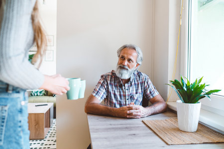 Senior man with grey hair and a beard sitting by the dining table while unrecognizable woman brings coffee to the tableの写真素材