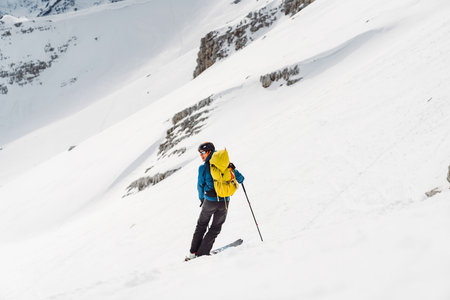 View from the back man ski touring with a yellow backpack, descending down the snowy mountainの写真素材