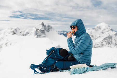 Mountaineer in blue jacket sitting on cold snow eating a sandwich on top of the mountainの写真素材