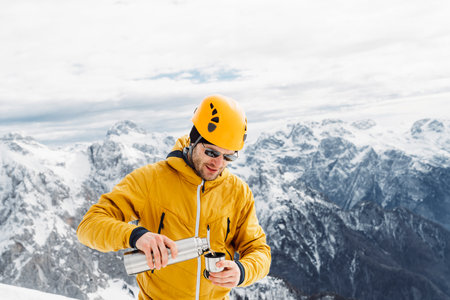 Mountaineer in a yellow jacket and helmet pouring himself some tea, view of snowy Alps in the backgroundの写真素材