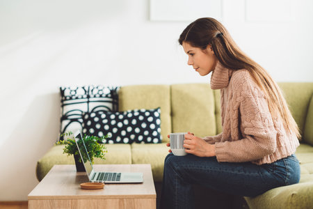 Side view of a woman sitting on the couch working on laptop while having her morning coffeeの写真素材