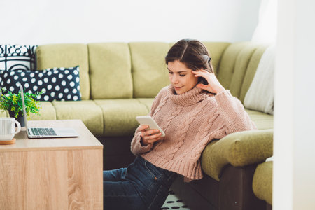 Young woman sitting on the carpet leaning on the couch while looking at her phoneの写真素材