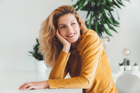 Waist up portrait curly blonde woman sitting by the table leaning her head on the palm of her hand, smiling while looking away from the cameraの写真素材