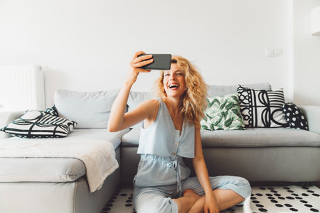 Cheerful curly blonde woman sitting on the carpet in front of the sofa taking selfiesの写真素材