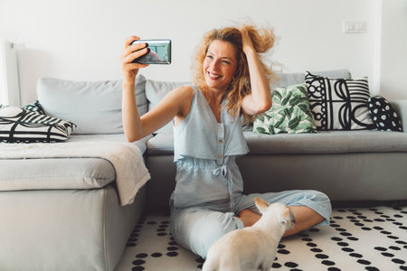 Smiling curly woman playing with her hair while taking selfies with her phone, sitting on the carpet by the couchの写真素材