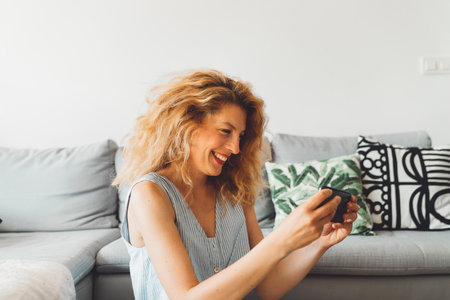 Side waist up portrait of a curly woman looking at her phone taking selfies while sitting down on the carpet in front of her sofaの写真素材