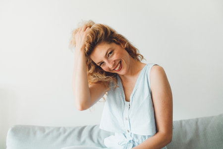 Waist up portait beautiful woman sitting on the couch playing with her hair smiling looking at the cameraの写真素材