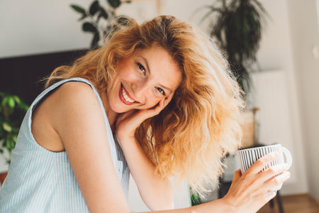 Cheerful curly woman looking at the camera with a big smile on her face wearing red lipstick and holding a cup of coffee in her handsの写真素材