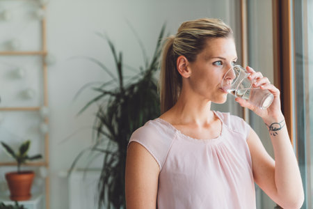 Portrait of blonde woman with a pony tail drinking a glass of water while standing by the window of her apartmentの写真素材