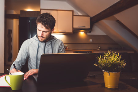 Caucasian man writing notes while attending evening online studying lessons on his laptop, sitting by the table with a cup of coffee next to the laptopの写真素材