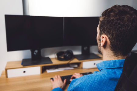 Over the shoulder view unrecognizable man in blue shirt working on two black screensの写真素材