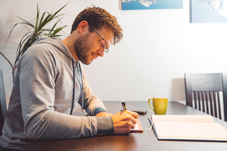 Side view of man with glasses wearing a tracksuit, sitting by the table making notes studying at homeの写真素材
