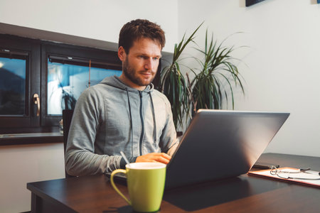 Adult bearded man studying online on his laptop sitting by the dining room table with a cup of coffee next to the laptopの写真素材