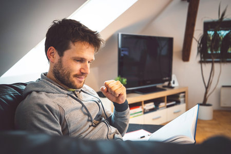 Waist up bearded adult man reading a notebook on the couch studying at homeの写真素材