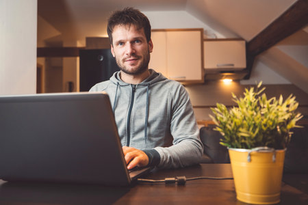 Front view of a man sitting by the table working on his laptop from home in the evening, looking at the cameraの写真素材