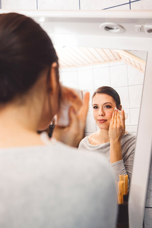 Over the shoulder view, young woman cleaning make up off her face standing in front of a mirrorの写真素材