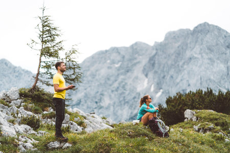 Couple of hikers taking a rest up in the mountains, woman sitting down on the grass and man looking up in the skyの写真素材