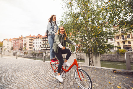 Sisters having a fun day in the city, riding a red bike, one of them standing on the bike, while the other rides itの写真素材