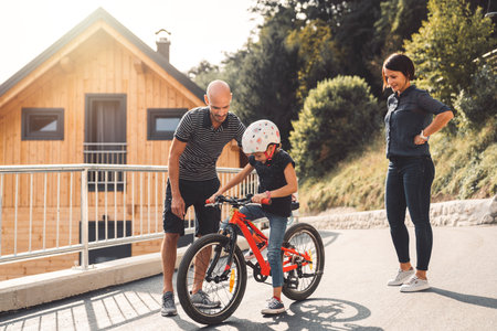 Dad teaching her young daughter how to ride a bike, while mom stands by looking at themの写真素材