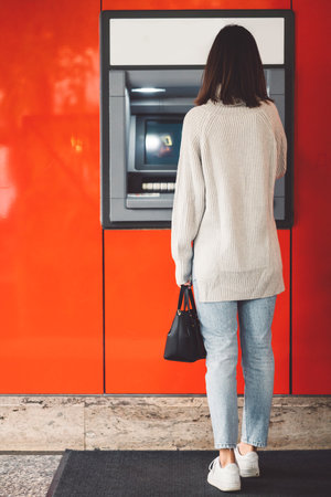 Vertical photo, back view of a woman standing in front of an ATM on a red wall, making a cash withdrawalの写真素材