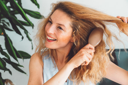 Waist up portrait smiling woman brushing her blonde curly hair with a hair brushの写真素材