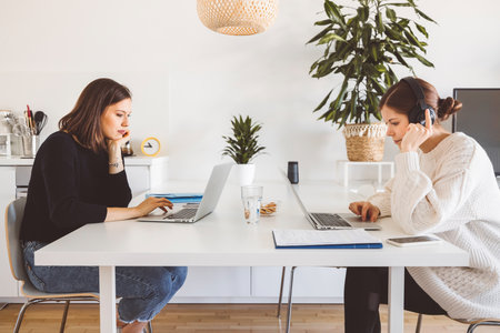 Side view of two young female students studying by the table each on her own laptop in a modern bright apartmentの写真素材