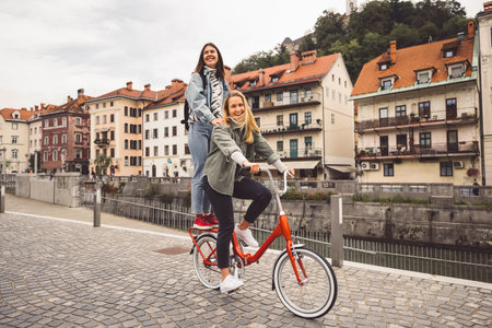 Couple of best friends riding a bike on the city streets, one of the women standing on the trunk of the bikeの写真素材