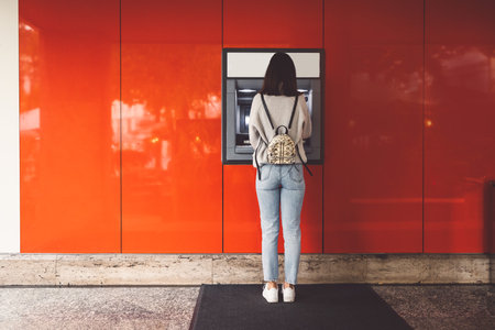Back view of young woman standing in front of an ATM machine on a red wallの写真素材