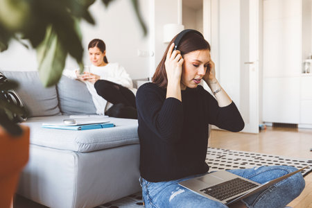 Two young women college students in their apartment studying on laptops, one in front putting head phones onの写真素材